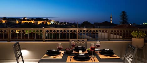 A rooftop terrace with a table set for two, overlooking a cityscape at dusk.