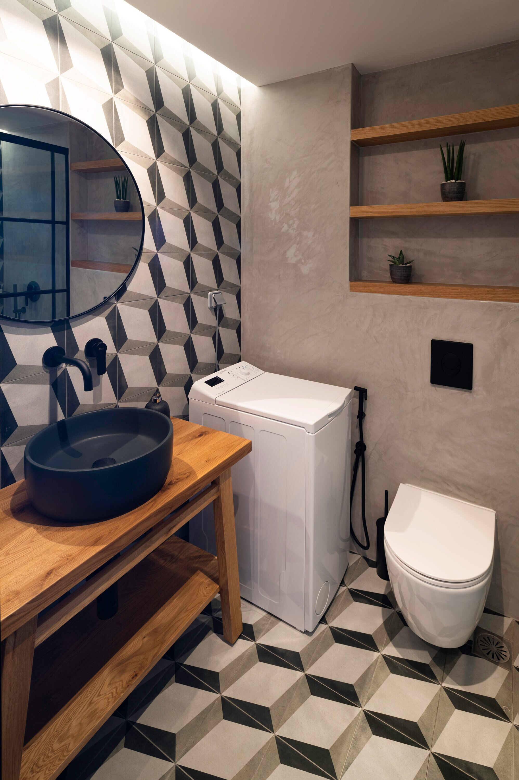 Un baño moderno con piso de baldosas de diseño geométrico, un espejo redondo, un tocador de madera con lavamanos negro, una lavadora blanca y un inodoro blanco.