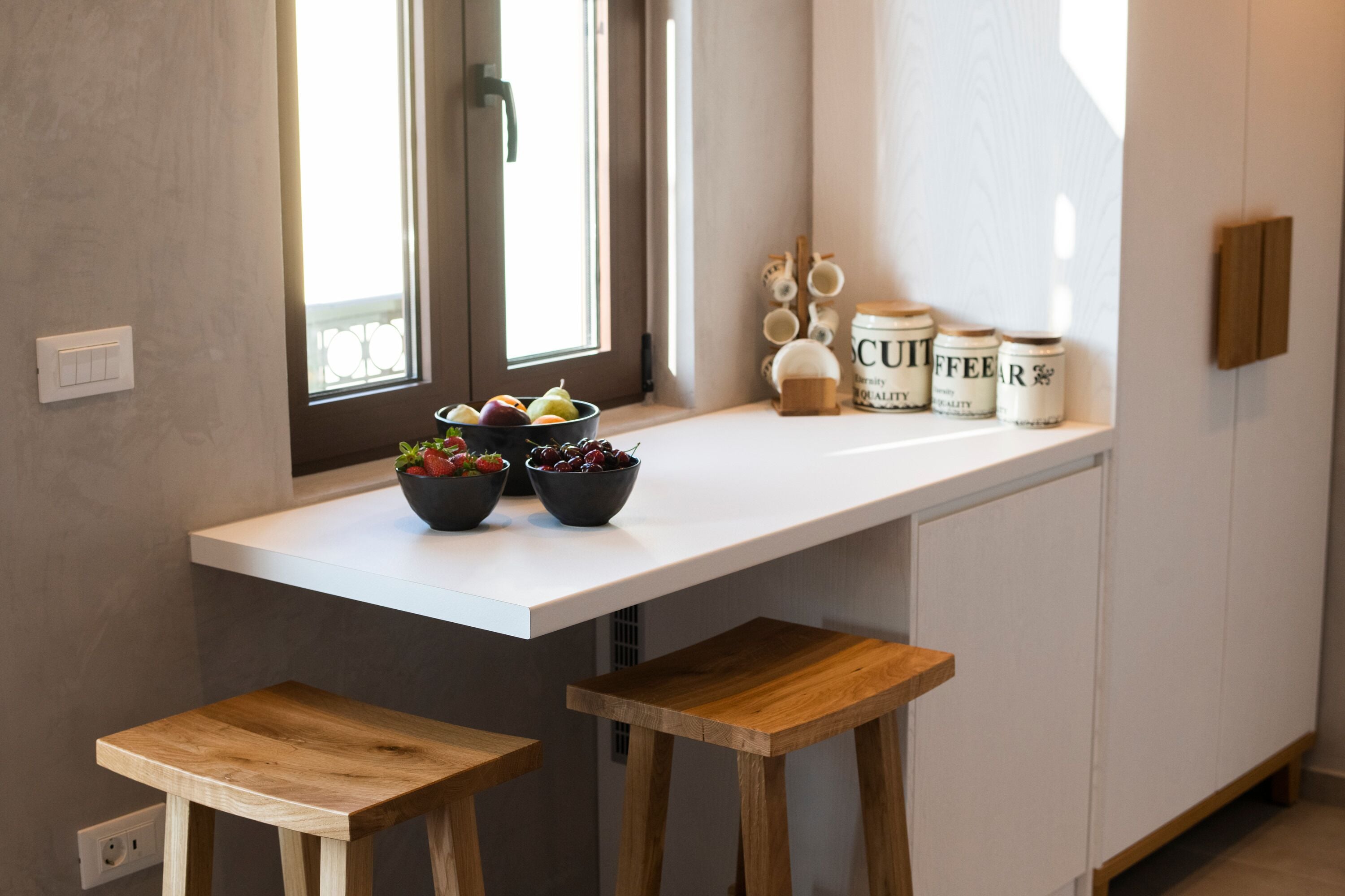 Una cocina con encimera blanca, dos taburetes de madera, un ventanal con cortinas y frascos etiquetados 'CUFFEE' y 'QUALITY'.