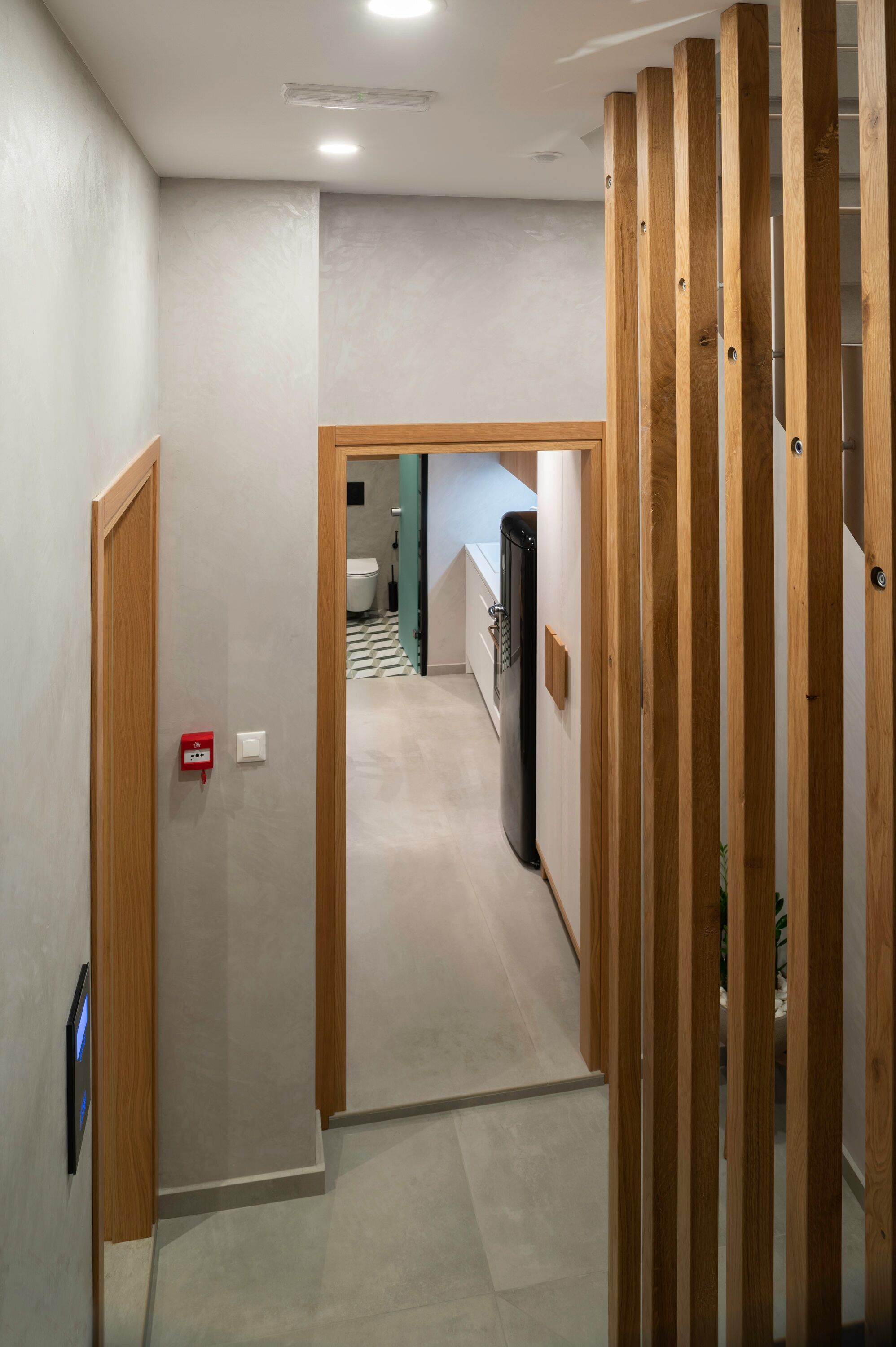 Un couloir avec une cloison en bois, une boîte rouge contenant du matériel de premiers secours, et on aperçoit une salle de bain avec des toilettes et un réfrigérateur.