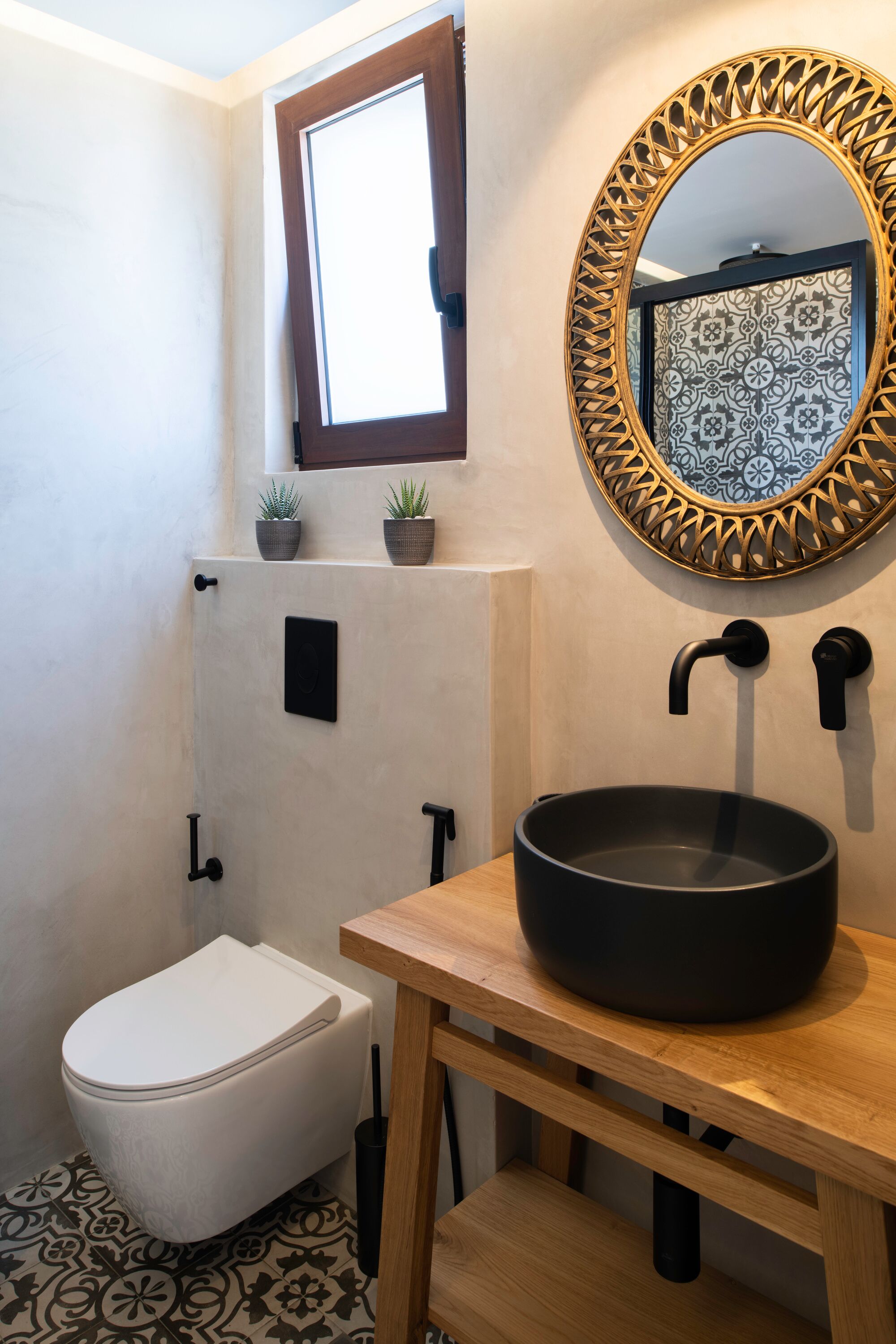 Un baño con inodoro blanco, lavamanos negro de forma redonda, espejo decorativo con marco estampado y una ventana sobre el inodoro.
