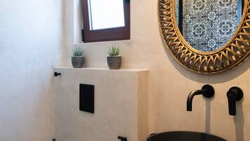 Un baño con inodoro blanco, lavamanos negro de forma redonda, espejo decorativo con marco estampado y una ventana sobre el inodoro.