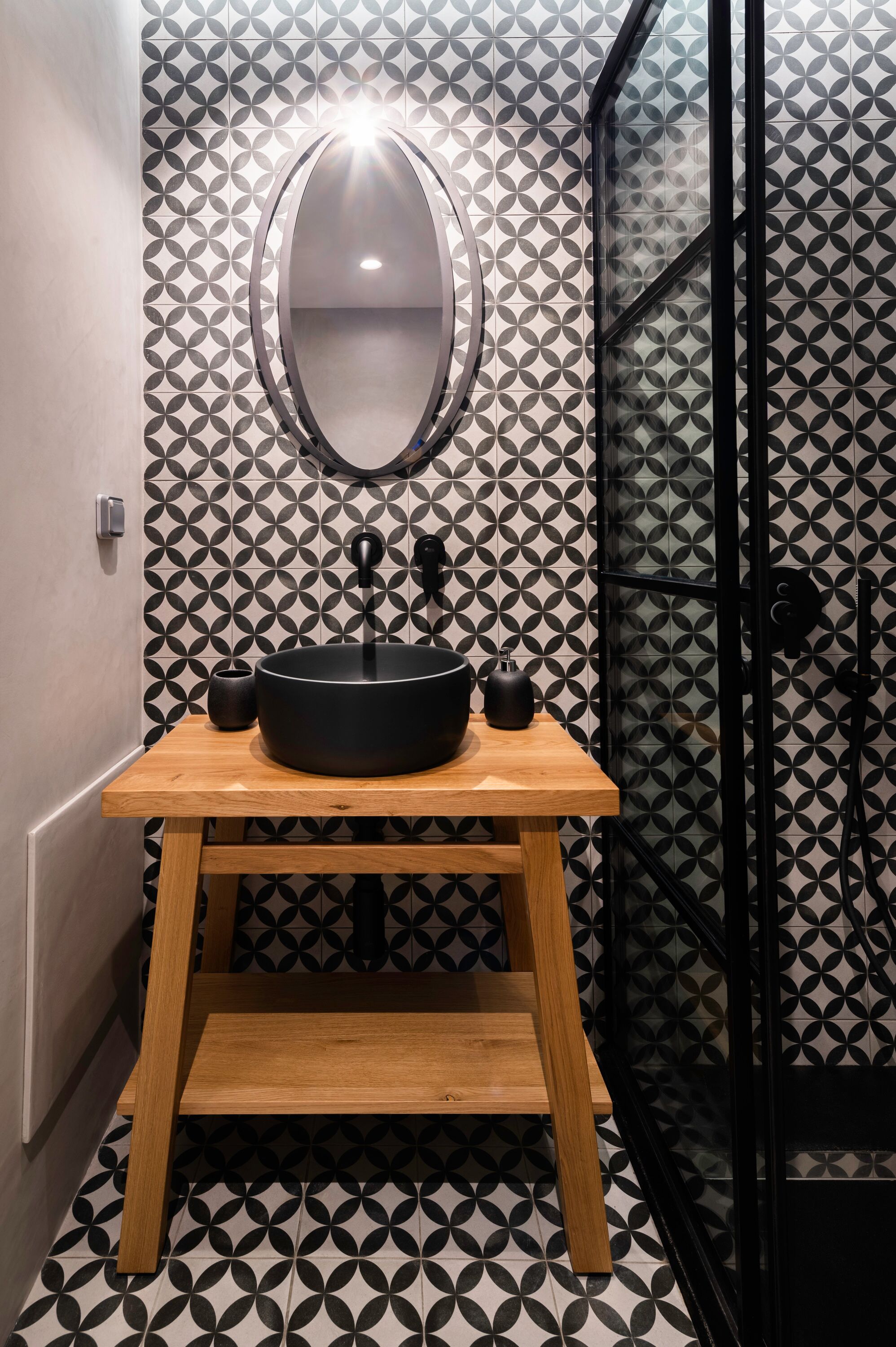 Un baño con un lavabo negro sobre un mueble de madera, un espejo redondo y una pared con estampado geométrico.