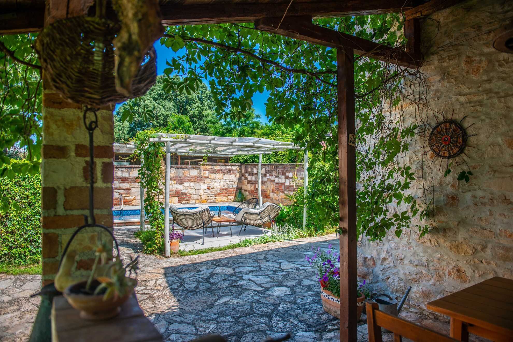 A stone patio with a pergola, outdoor seating, and a view of a pool area.