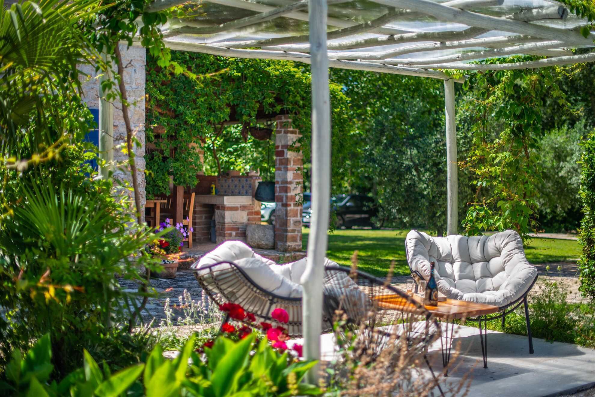 A covered patio with two cushioned chairs and a small table, surrounded by lush greenery and a brick structure.