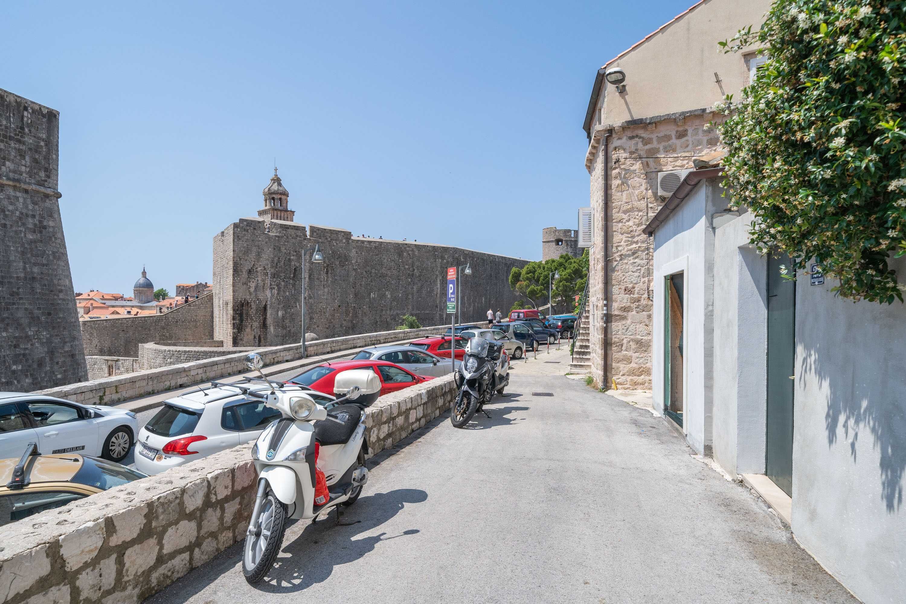 Un espace de stationnement avec des voitures et des motos, un mur de pierre et, au fond, un bâtiment doté d’un dôme et d’un clocher.