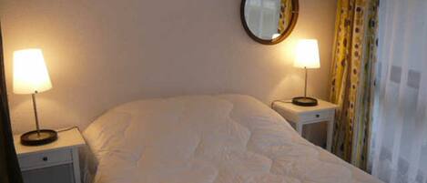 A neatly made bed with a white duvet, two bedside tables with lamps, a round mirror on the wall, and a curtain on the window.