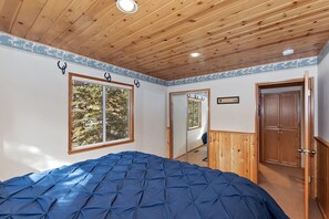 Ein Schlafzimmer mit einem großen Bett, einer Holzbalkendecke, einem Fenster mit Blick auf Bäume und einer Tür zu einem anderen Raum.