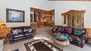 A spacious living room with two leather sofas, a glass coffee table, and a wooden ceiling. A framed picture hangs on the wall, and a staircase leads to a kitchen area. - Falling Leaves by Destination Big Bear : Premiere Moonridge Cabin Offering a Perfect Setting for Escaping to the Mountains! (Big Bear Lake)