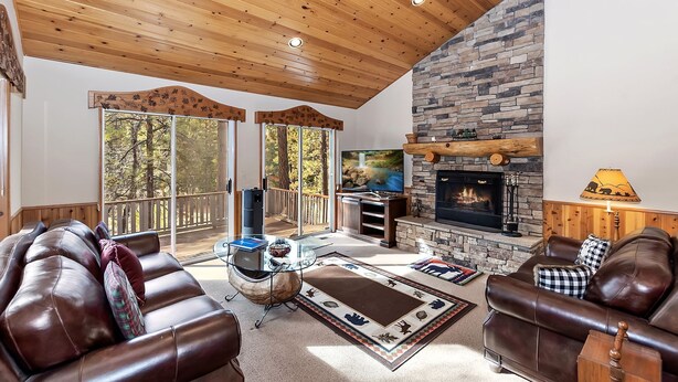 A cozy living room with a stone fireplace, leather sofas, a glass coffee table, and a large window overlooking a forest. - Falling Leaves by Destination Big Bear : Premiere Moonridge Cabin Offering a Perfect Setting for Escaping to the Mountains! (Big Bear Lake)