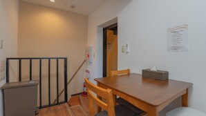 A small, clean room with a wooden table, a chair, a desk, and a trash bin. There is a staircase with a handrail and a fire extinguisher on the wall.
