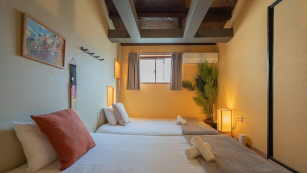 A hotel room with two beds, a large window, a potted plant, and a painting on the wall.