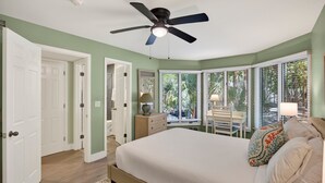 Une chambre à coucher avec un lit, un ventilateur de plafond, une fenêtre avec des stores, une table de chevet et une chaise près de la fenêtre.