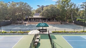 Un court de tennis doté d’un auvent vert, d’un espace pour s’asseoir et d’arbres en arrière-plan.