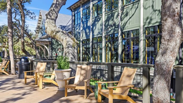 Une terrasse en bois, agrémentée de chaises et d’une table, entourée d’arbres et bordée d’un bâtiment doté de grandes fenêtres.