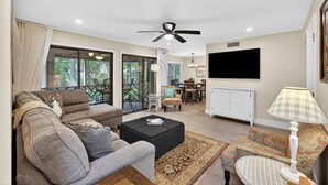 Un salon comprenant un canapé, un pouf et un fauteuil, un coin repas, un ventilateur de plafond, un téléviseur et une fenêtre donnant sur un paysage verdoyant.