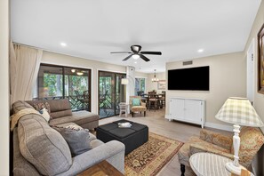 A living room with a sofa, ottoman, and armchair, a dining area, a ceiling fan, a TV, and a window with a view of greenery.
