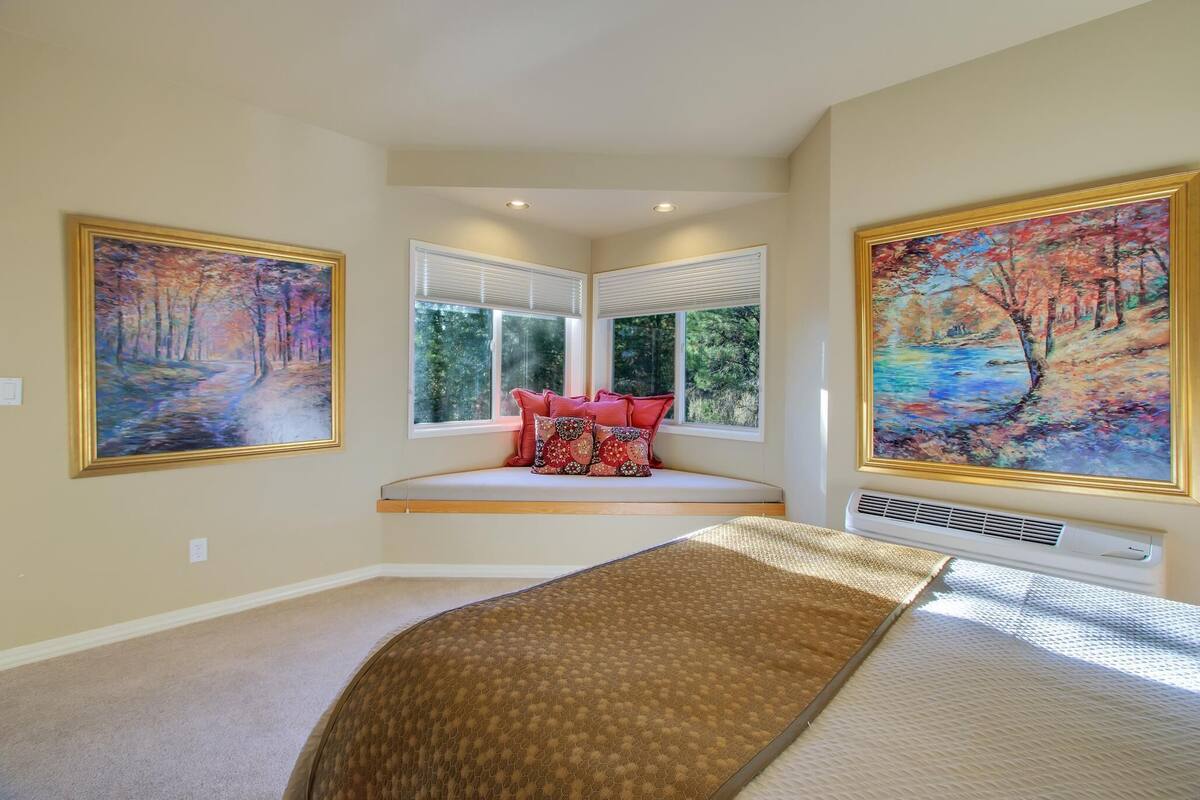 A bedroom with a large bed, a window seat with red pillows, and two framed paintings on the walls.