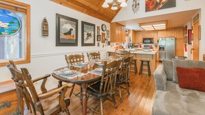 A dining area with wooden chairs and a table set for six, a kitchen with wooden cabinets, and a gray sofa with a red pillow. - BlackDog Hideaway_Hot Tub, (2) Arcades, Foosball, PingPong, Smart Tv's, Cable + HOA Amenity Passes (Truckee)