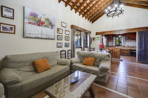 A living room with two sofas, a glass coffee table, and a kitchen area visible through an open door.