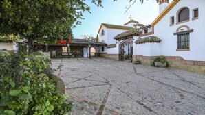 A courtyard with cobblestone flooring, a white building with a tiled roof, and a tree with red flowers. - Holiday Home ‘La Cazuela’ with Mountain View, Pool, Garden & Terraces (Arcos de la Frontera)