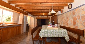 Une cuisine rustique avec des poutres en bois, un mur en pierre, une table à manger recouverte d’une nappe verte et un banc.