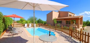 A swimming pool with lounge chairs and a table, surrounded by a wooden fence and greenery.