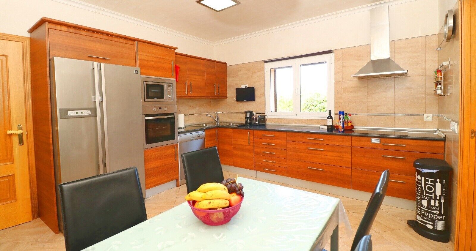 A modern kitchen with wooden cabinets, stainless steel appliances, a dining table with a fruit bowl, and a window providing natural light.