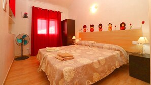 A bedroom with a bed, red curtains, a bedside table with a lamp, a wall-mounted shelf with decorative items, and a standing fan.