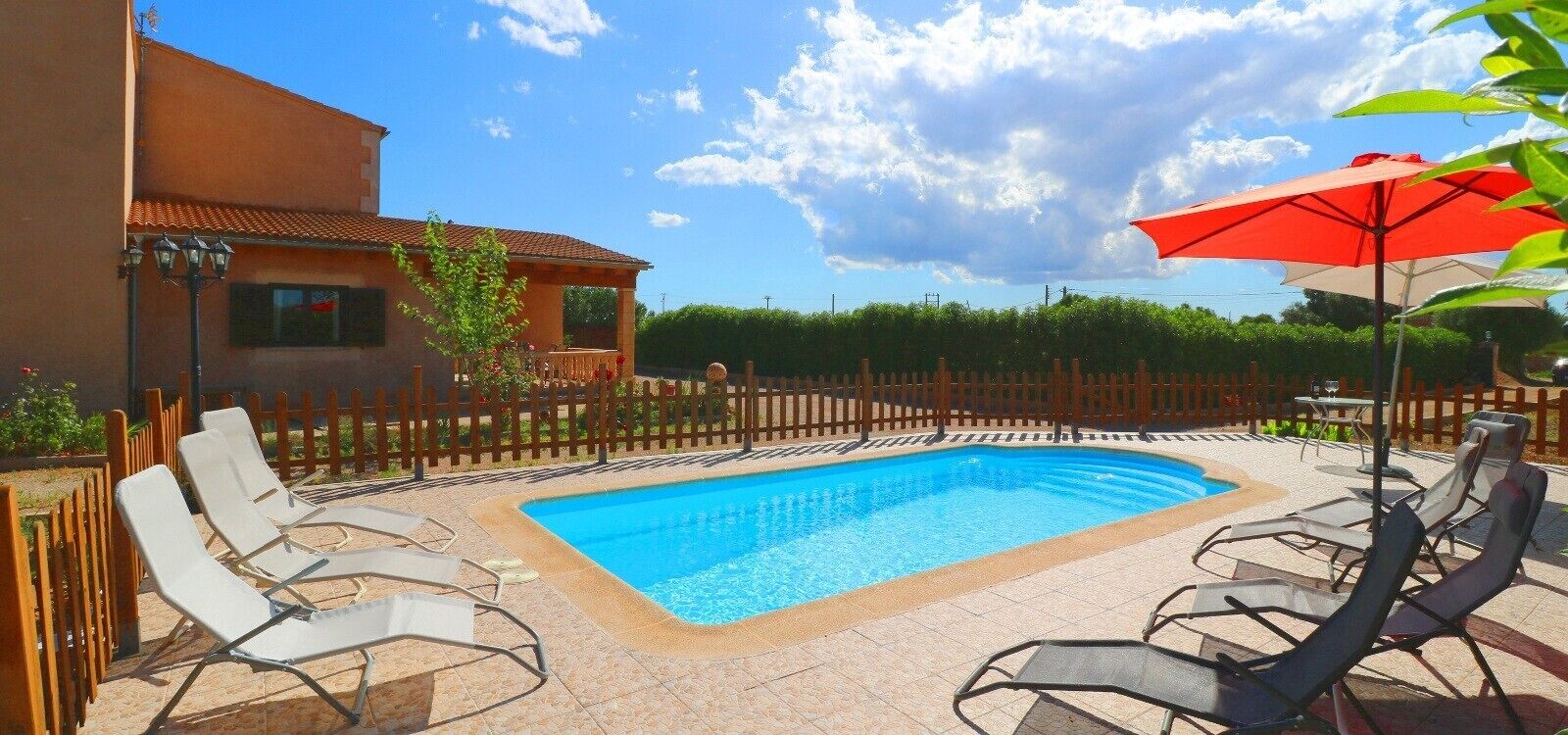 Une piscine avec des chaises longues et un parasol rouge, entourée d’une clôture en bois et d’un bâtiment.