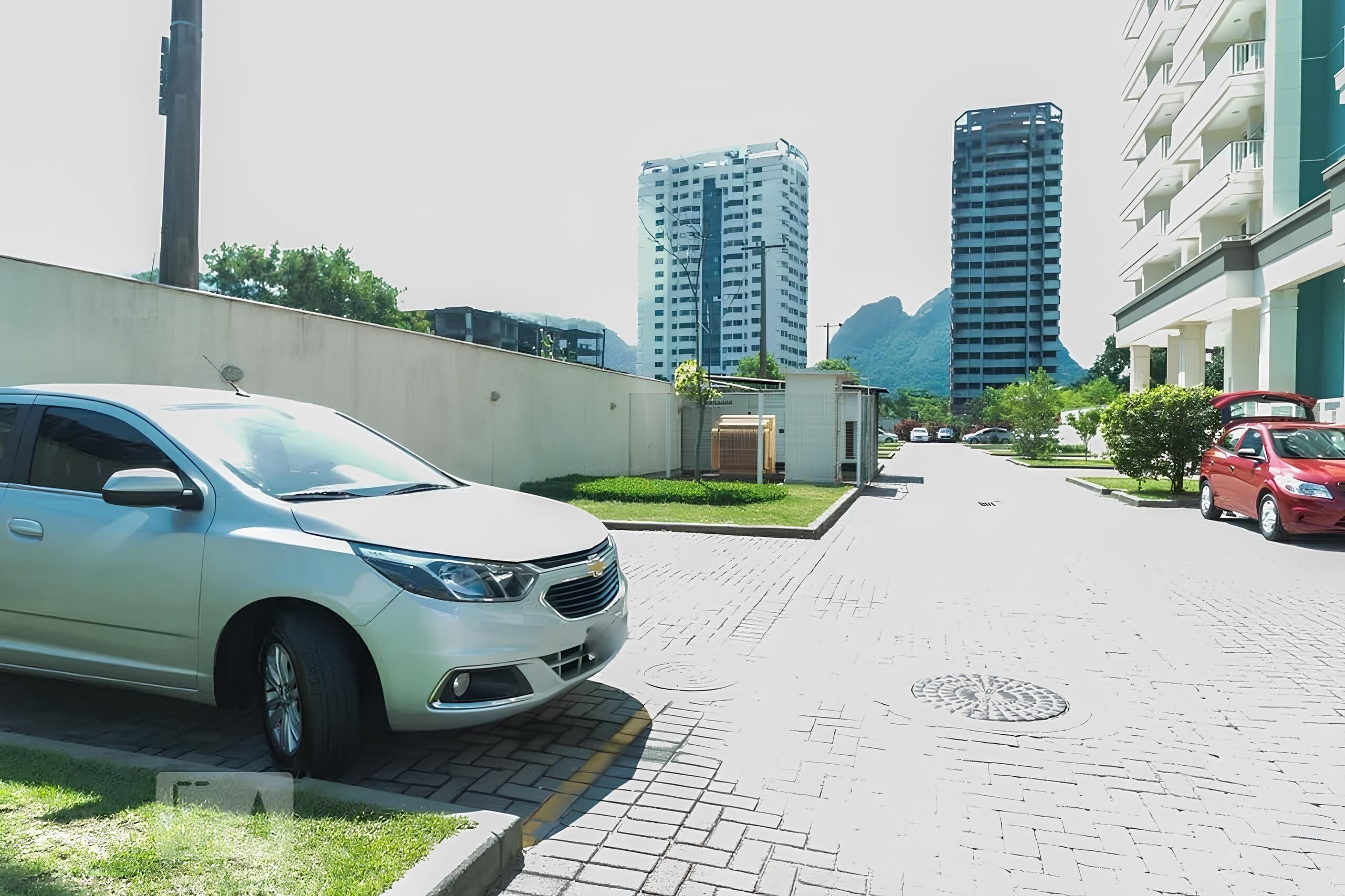 Uma rua residencial com carros estacionados, um carro branco em primeiro plano e edifícios altos ao fundo.