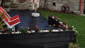 En byggnad av sten med en röd dörr och ett tak täckt med tegelpannor, ett vitt parasoll och blomrabatter på gården.