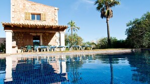 Una piscina con agua azul cristalina, un edificio de piedra con balcón y palmeras al fondo.