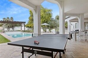 Une table de ping-pong installée à l’extérieur, avec une planche de skate à proximité, le tout entouré de mobilier de jardin et d’une piscine.