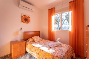 Un dormitorio con cama, mesita de noche, lámpara, ventana con cortinas y aire acondicionado instalado en la pared.