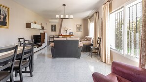 Ein modernes Wohnzimmer mit einer Couch, einem Esstisch, Stühlen, einem Fernseher und einem großen Fenster mit Vorhängen.