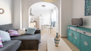 Una sala de estar con un sofá, una mesa de centro con un jarrón, un televisor en un mueble bajo y un arco que conduce a una cocina y comedor.