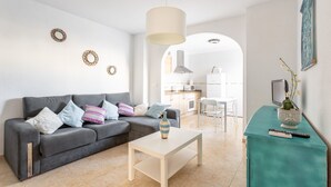 Una sala de estar con un sofá gris, una mesa de centro blanca, una mesita auxiliar color turquesa y una cocina al fondo.
