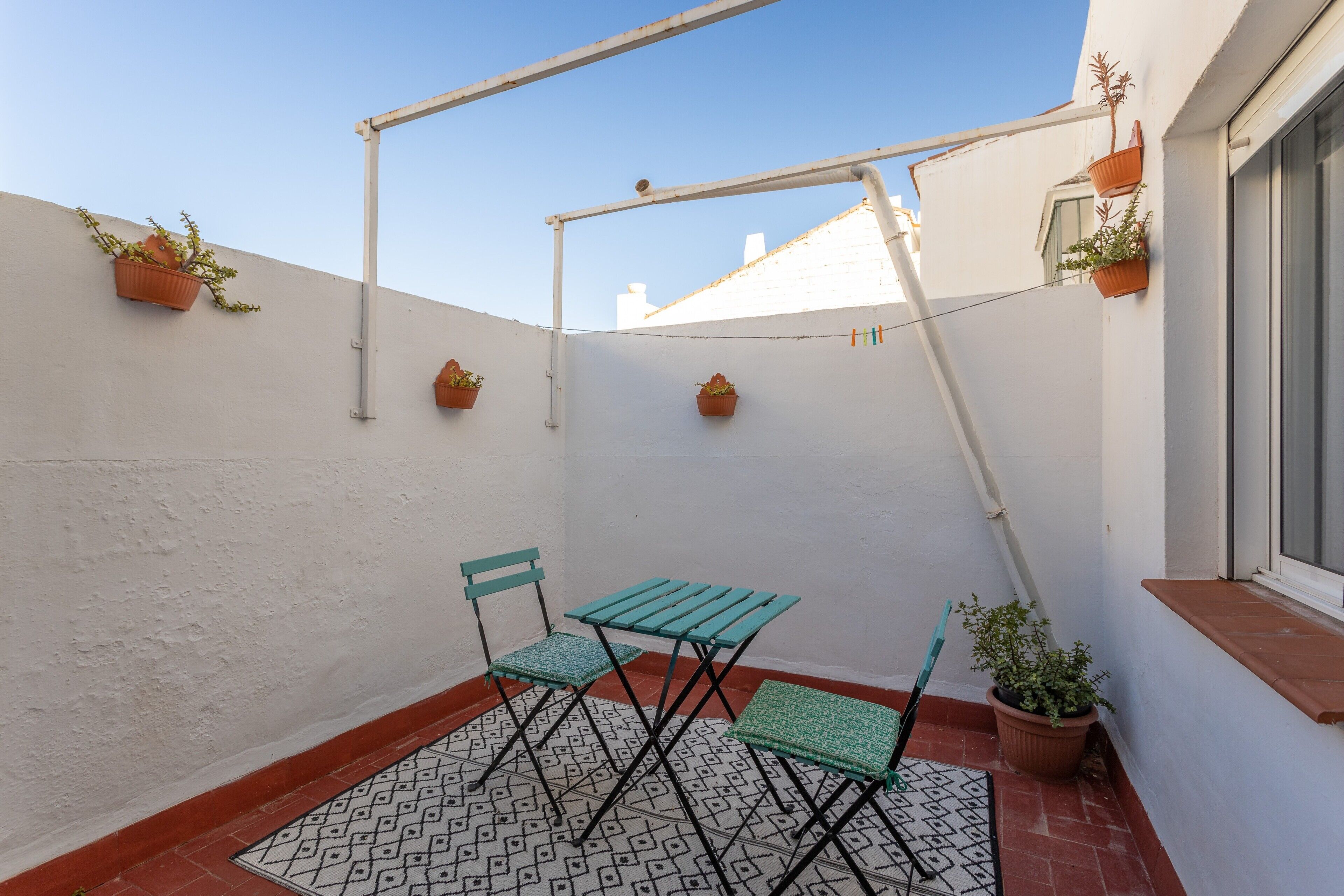 A small outdoor patio with a table and chairs, potted plants, and a white wall.