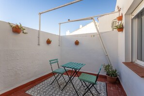 Een klein terras buiten met een tafel, stoelen, potplanten en een witte muur.