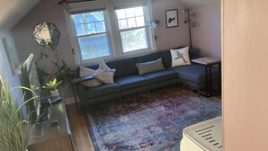 Uma sala de estar aconchegante com um sofá cinza, almofadas estampadas, uma janela grande com persianas, um tapete decorativo e uma pequena mesa com uma planta.