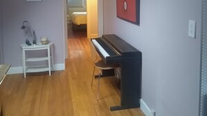 Um quarto com um piano, uma mesa branca com uma estatueta de gato e uma porta que dá para outro cômodo.