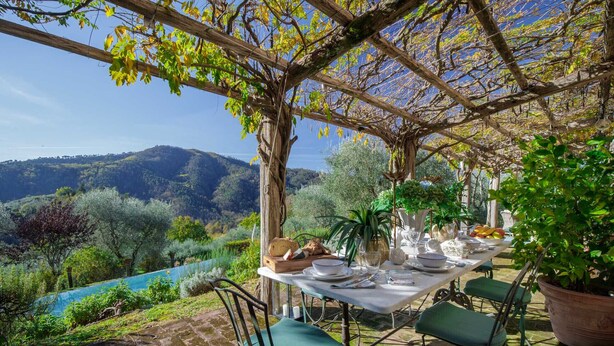 An outdoor dining area with a table set for four, surrounded by greenery and a view of hills.