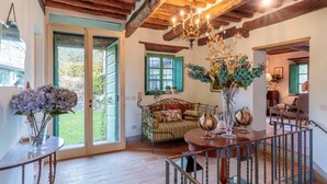 A spacious room with a wooden ceiling, a sofa with patterned cushions, a table with decorative items, and a large window with green shutters.