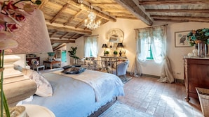 A rustic bedroom with exposed wooden beams, a chandelier, a bed with a blue cover, a desk with lamps, and a mirror.
