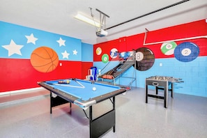 Una mesa de billar con superficie de fieltro azul, una mesa de fútbol de mesa y una pared con dianas circulares de colores.