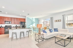 A modern kitchen with stainless steel appliances, a dining area with a white table and chairs, a living room with a white sofa and a coffee table, and a large window with curtains.
