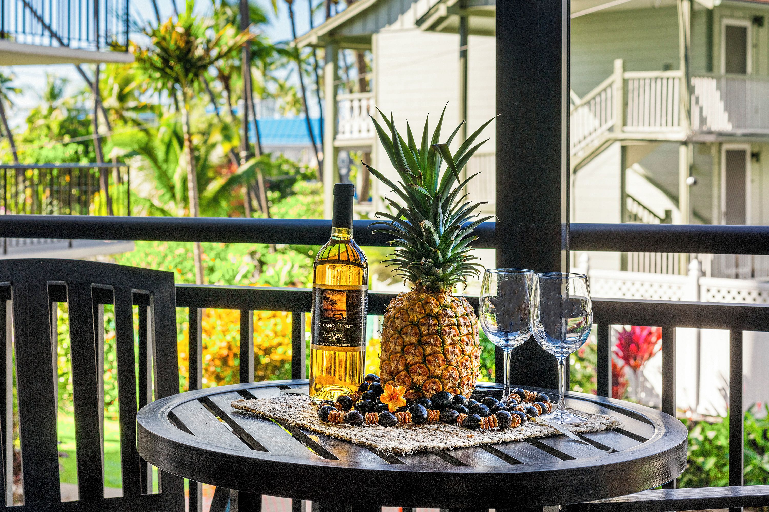 A table with a bottle of alcohol, a pineapple, and two glasses on a balcony overlooking a tropical garden and a building with balconies.