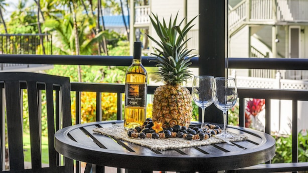 Ein Tisch mit einer Alkoholflasche, einer Ananas und zwei Gläsern auf einem Balkon mit Blick auf einen tropischen Garten und ein Gebäude mit Balkonen.