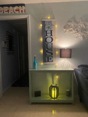 Et værelse med et vægmonteret skilt med teksten "THE HOUSE", vægkunst i form af en dekorativ søstjerne, et lille bord med en blå flaske og en geometrisk lampe, samt en tændt lygte.